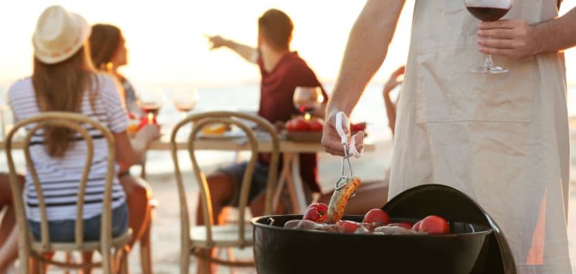 person grilling shrimp on a beach barbecue while people drink wine at a table