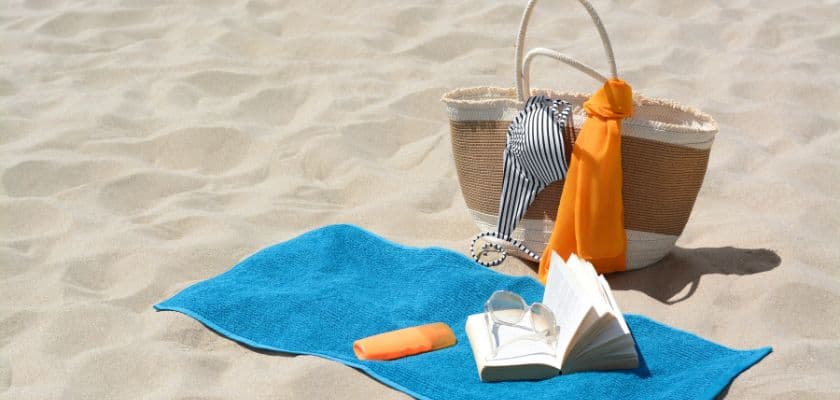 A blue towel lies on the sand with an open book, sunglasses, sunscreen, and a beach bag holding a striped swimsuit and an orange towel.